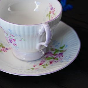 May include: A white porcelain teacup and saucer set featuring a delicate ribbed texture and a hand-painted floral pattern in shades of pink, purple, and green. The rim of both the cup and saucer is accented with a thin, soft purple painted line.