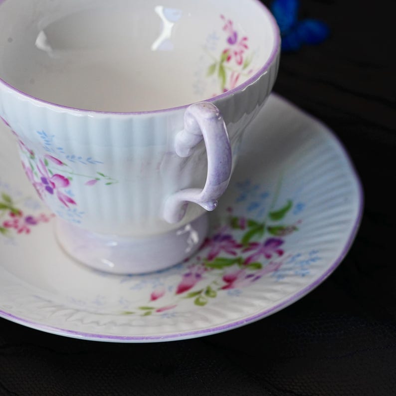 May include: A white porcelain teacup and saucer set featuring a delicate ribbed texture and a hand-painted floral pattern in shades of pink, purple, and green. The rim of both the cup and saucer is accented with a thin, soft purple painted line.
