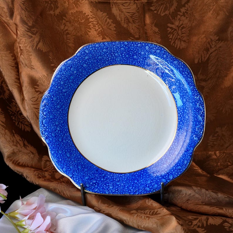 May include: A white plate with a blue patterned rim and a gold trim. The rim has a scalloped edge. The plate is set against a brown, textured background. The plate is a decorative item.