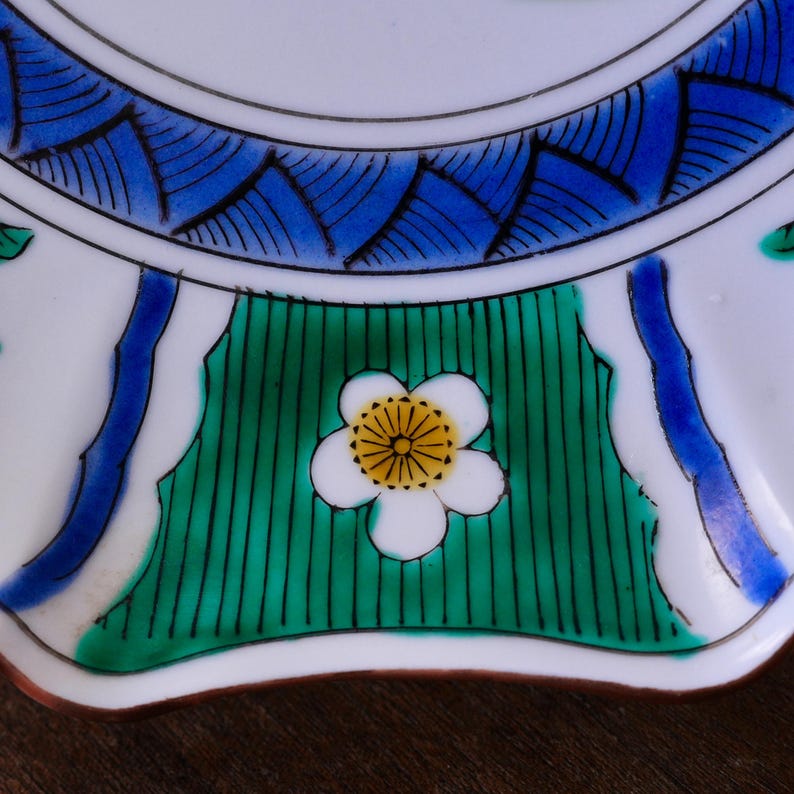 May include: Close-up of a white ceramic plate with a floral design. The plate features a white flower with a yellow centre, set against a green background with vertical black lines. The rim has blue and white patterns.