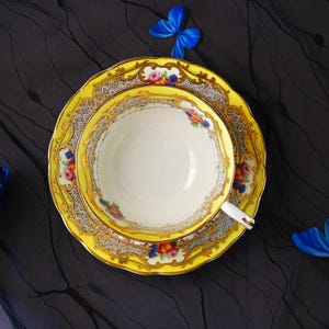 Peut inclure: Un ensemble tasse et soucoupe en porcelaine avec une bordure jaune vif orn&eacute;e de filigrane dor&eacute;, un motif en treillis gris et de petites grappes de fleurs roses, bleues et orange. L'int&eacute;rieur de la tasse est blanc et l'anse est blanche et dor&eacute;e.