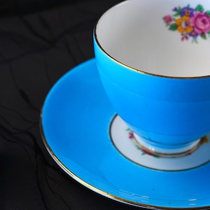 Op de afbeelding: Een fel turquoise theekop en schotel set met een goudkleurige rand. De binnenkant van de kop en het midden van de schotel zijn versierd met een klein, kleurrijk bloemenpatroon bestaande uit roze, gele en paarse bloemen met groene bladeren.