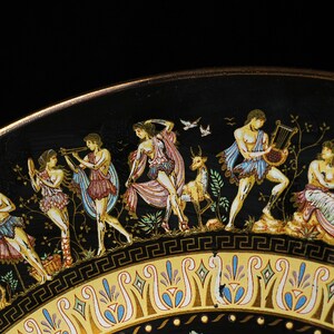 May include: A decorative black plate with a gold rim and a detailed frieze depicting classical figures in colorful attire. The figures are engaged in various activities, including playing instruments and dancing. The plate's border features a repeating geometric pattern.