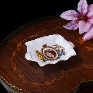 May include: A white porcelain dish with a scalloped edge and gold trim, featuring a central portrait of Queen Elizabeth II surrounded by a royal crest with a lion and unicorn, and the text June 2nd 1953 inscribed at the base of the emblem.