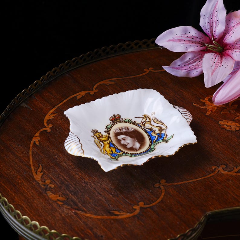 May include: A white porcelain dish with a scalloped edge and gold trim, featuring a central portrait of Queen Elizabeth II surrounded by a royal crest with a lion and unicorn, and the text June 2nd 1953 inscribed at the base of the emblem.