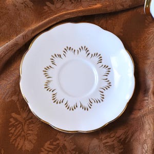 May include: A white porcelain saucer with a scalloped edge and gold trim. The saucer features a gold leaf-like pattern around the center. This elegant saucer is perfect for serving tea or coffee and adds a touch of sophistication to any table setting.