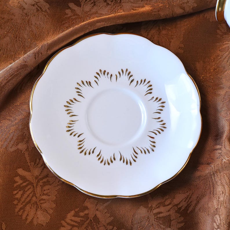 May include: A white porcelain saucer with a scalloped edge and gold trim. The saucer features a gold leaf-like pattern around the center. This elegant saucer is perfect for serving tea or coffee and adds a touch of sophistication to any table setting.
