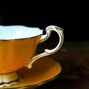 May include: A close-up shot of a vintage teacup and saucer. The cup and saucer are a warm yellow-orange with gold trim. The handle is white with gold detailing. The cup's interior is a pale blue.
