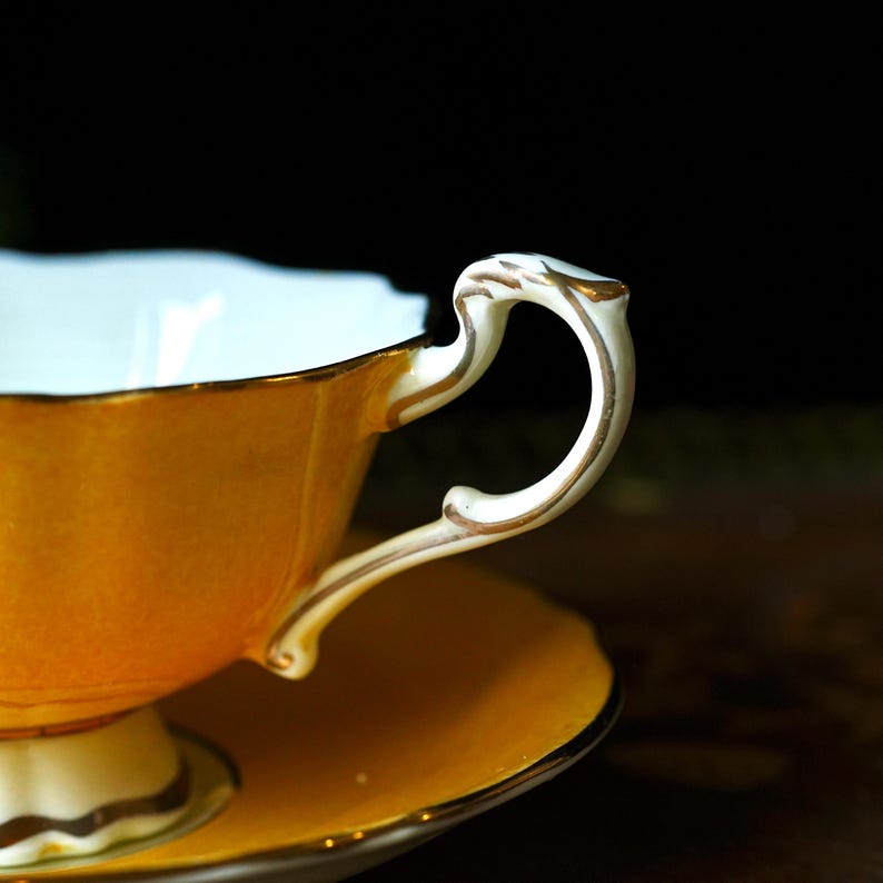 May include: A close-up shot of a vintage teacup and saucer. The cup and saucer are a warm yellow-orange with gold trim. The handle is white with gold detailing. The cup's interior is a pale blue.