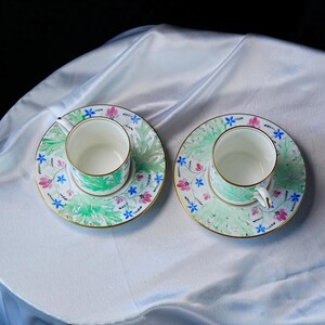 May include: Two white porcelain teacups and matching saucers featuring a delicate hand-painted pattern of green foliage, small pink flowers, and tiny blue blossoms. Each piece is finished with a thin gold rim along the edges and handles.