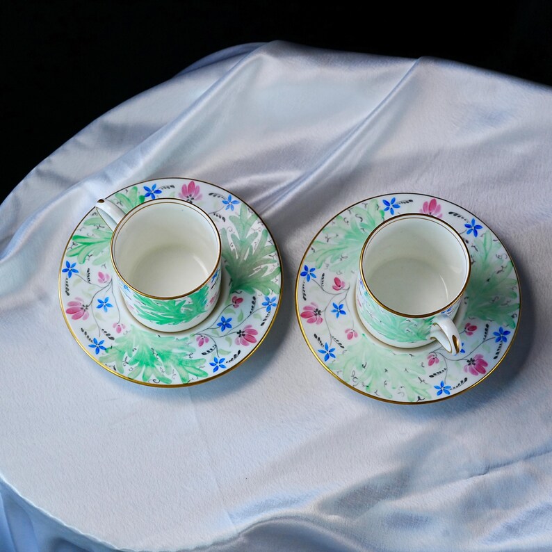 May include: Two white porcelain teacups and matching saucers featuring a delicate hand-painted pattern of green foliage, small pink flowers, and tiny blue blossoms. Each piece is finished with a thin gold rim along the edges and handles.