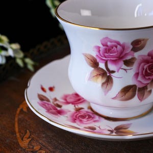 Op de afbeelding: Witte theekop en schotel set met roze rozen bloemenpatroon en gouden rand. De theekop heeft een gebogen vorm en de schotel is rond. De rozen staan in volle bloei met bruine bladeren. De set staat op een houten oppervlak.