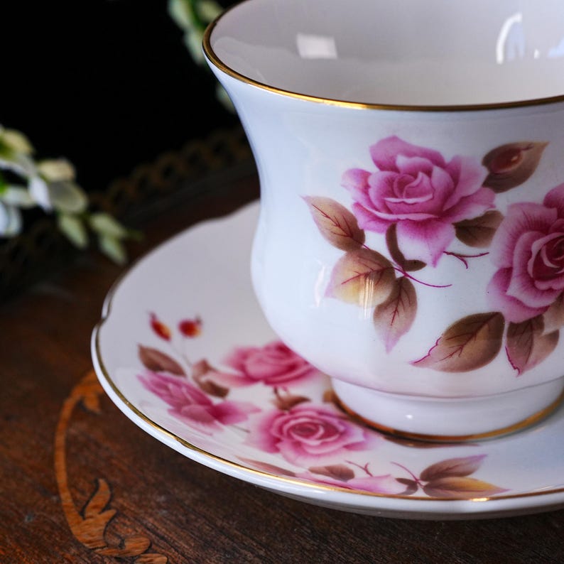 Op de afbeelding: Witte theekop en schotel set met roze rozen bloemenpatroon en gouden rand. De theekop heeft een gebogen vorm en de schotel is rond. De rozen staan in volle bloei met bruine bladeren. De set staat op een houten oppervlak.
