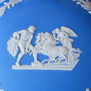May include: A bright blue ceramic surface featuring a raised white relief decoration. The design depicts a figure standing beside a chariot pulled by two horses, with a winged figure seated in the chariot. A leafy border pattern is visible at the top.