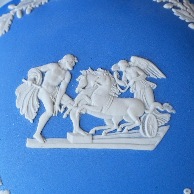 May include: A bright blue ceramic surface featuring a raised white relief decoration. The design depicts a figure standing beside a chariot pulled by two horses, with a winged figure seated in the chariot. A leafy border pattern is visible at the top.