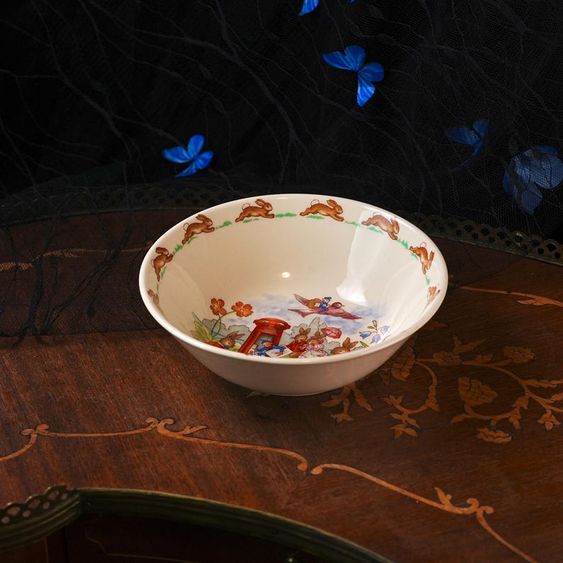 May include: A cream-colored ceramic bowl featuring a repeating pattern of brown rabbits along the inner rim. The center of the bowl displays a colorful illustration of rabbits, flowers, and a red telephone booth under a blue sky.