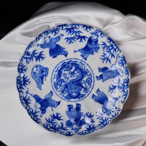 Puede incluir: Plato de cer&aacute;mica blanca con borde festoneado, con ilustraciones azules. El centro muestra un drag&oacute;n rodeado por un borde detallado. Figuras con vestimenta tradicional y ramitas de bamb&uacute; adornan la superficie del plato, creando un dise&ntilde;o cl&aacute;sico de inspiraci&oacute;n asi&aacute;tica.