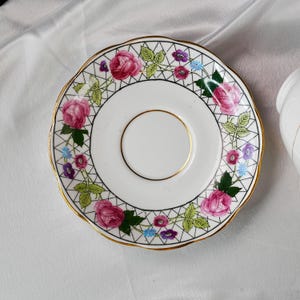 Op de afbeelding: Witte keramische schotel met een gouden rand en een bloemenontwerp. De schotel heeft roze rozen, paarse en blauwe bloemen, met groene bladeren en wijnstokken. De rand heeft een zwart geometrisch patroon.