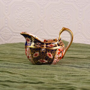 May include: A decorative porcelain creamer with a gold handle and spout. The creamer features a detailed floral pattern in red, white, and blue, with gold accents. The creamer is set against a green cloth background.
