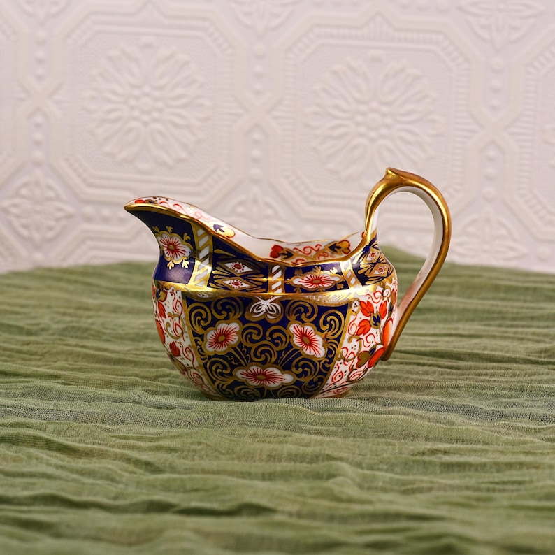 May include: A decorative porcelain creamer with a gold handle and spout. The creamer features a detailed floral pattern in red, white, and blue, with gold accents. The creamer is set against a green cloth background.
