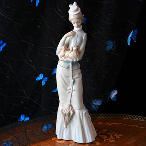 K&ouml;nnte beinhalten: Eine Porzellanfigur einer Person in einer hellblauen Jacke und einem langen wei&szlig;en Rock, die einen kleinen Hund im Arm h&auml;lt. Die Figur tr&auml;gt einen kleinen Hut und h&auml;lt einen geschlossenen Regenschirm mit einem hellrosa Schirmdach.