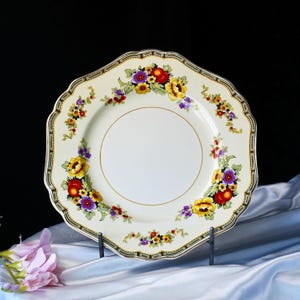 May include: An off-white plate with a scalloped edge and a floral design. The plate has a gold rim and a border with a black, green, and gold pattern. The floral design includes red, yellow, and purple flowers.