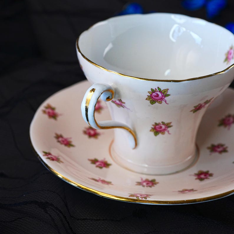 Może przedstawiać: Biały porcelanowy zestaw filiżanki ze spodkiem, ozdobiony delikatnym wzorem małych r&oacute;żowych r&oacute;ż z zielonymi liśćmi. Krawędź filiżanki i spodka wykończona jest złotem. Uchwyt filiżanki posiada r&oacute;wnież złotą linię akcentującą.