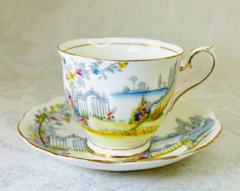 Può includere: Un set vintage di tazza da tè e piattino con un delicato motivo floreale. La tazza e il piattino presentano una scena di giardino dipinta a mano con un cancello, alberi e fiori in tonalità di blu, giallo e rosa. Il bordo della tazza e del piattino è rifinito con una sottile fascia dorata.