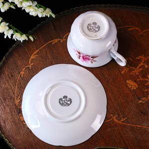 Op de afbeelding: Een witte Royal Stafford theekop en schotel met roze rozen en gouden rand. De omgedraaide kop toont het logo. De set staat op een decoratief houten oppervlak.