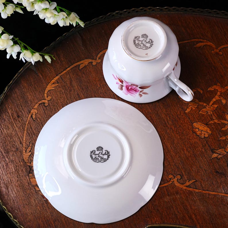 Op de afbeelding: Een witte Royal Stafford theekop en schotel met roze rozen en gouden rand. De omgedraaide kop toont het logo. De set staat op een decoratief houten oppervlak.