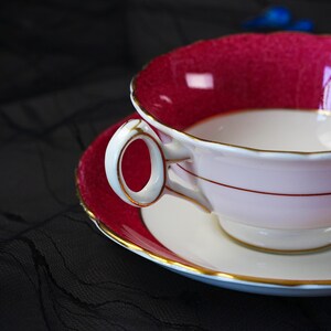 K&ouml;nnte beinhalten: Ein Porzellan-Tassenset mit Untertasse, das sich durch ein tief magentafarbenes Inneres und einen Rand mit strukturierter Oberfl&auml;che auszeichnet. Die Au&szlig;enseite der Tasse und die Mitte der Untertasse sind wei&szlig; und mit Goldrand verziert.