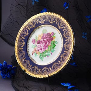 May include: A round porcelain plate featuring a central white basin decorated with a cluster of pink, purple, and yellow roses with green leaves. The wide rim is deep blue with an intricate gold filigree pattern and a scalloped gold edge.