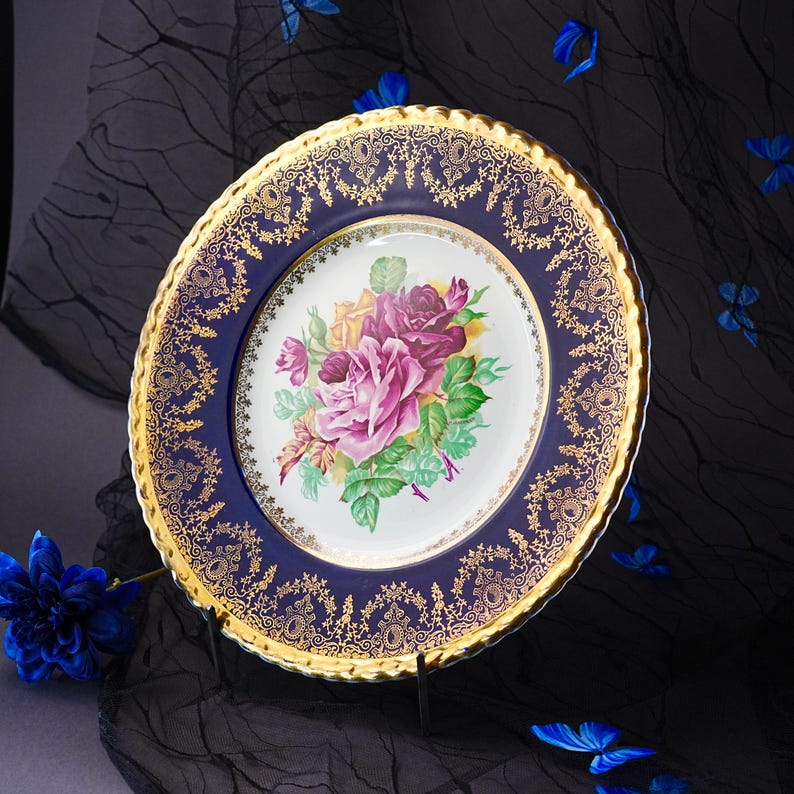 May include: A round porcelain plate featuring a central white basin decorated with a cluster of pink, purple, and yellow roses with green leaves. The wide rim is deep blue with an intricate gold filigree pattern and a scalloped gold edge.