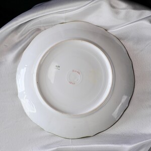 May include: White ceramic plate with a scalloped edge. The underside features a central red stamp with Japanese characters and the number 1817. A small square stamp is also visible. The plate is set against a white fabric background.