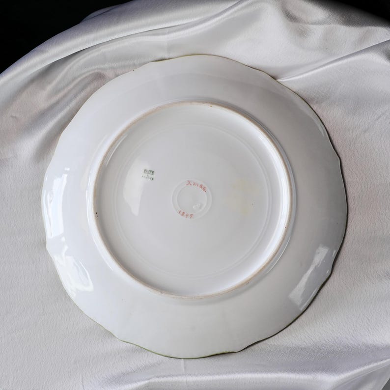 May include: White ceramic plate with a scalloped edge. The underside features a central red stamp with Japanese characters and the number 1817. A small square stamp is also visible. The plate is set against a white fabric background.