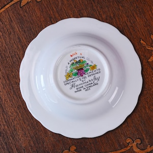 Puede incluir: Un platillo de porcelana blanca con borde festoneado. La parte inferior presenta un escudo floral colorido y texto negro que dice A PERPETUAL MEMENTO CORONATION SPECIALLY DESIGNED BY Hammersley BONE CHINA MADE IN ENGLAND 1953 con las iniciales ER.