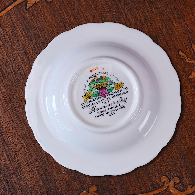 Puede incluir: Un platillo de porcelana blanca con borde festoneado. La parte inferior presenta un escudo floral colorido y texto negro que dice A PERPETUAL MEMENTO CORONATION SPECIALLY DESIGNED BY Hammersley BONE CHINA MADE IN ENGLAND 1953 con las iniciales ER.