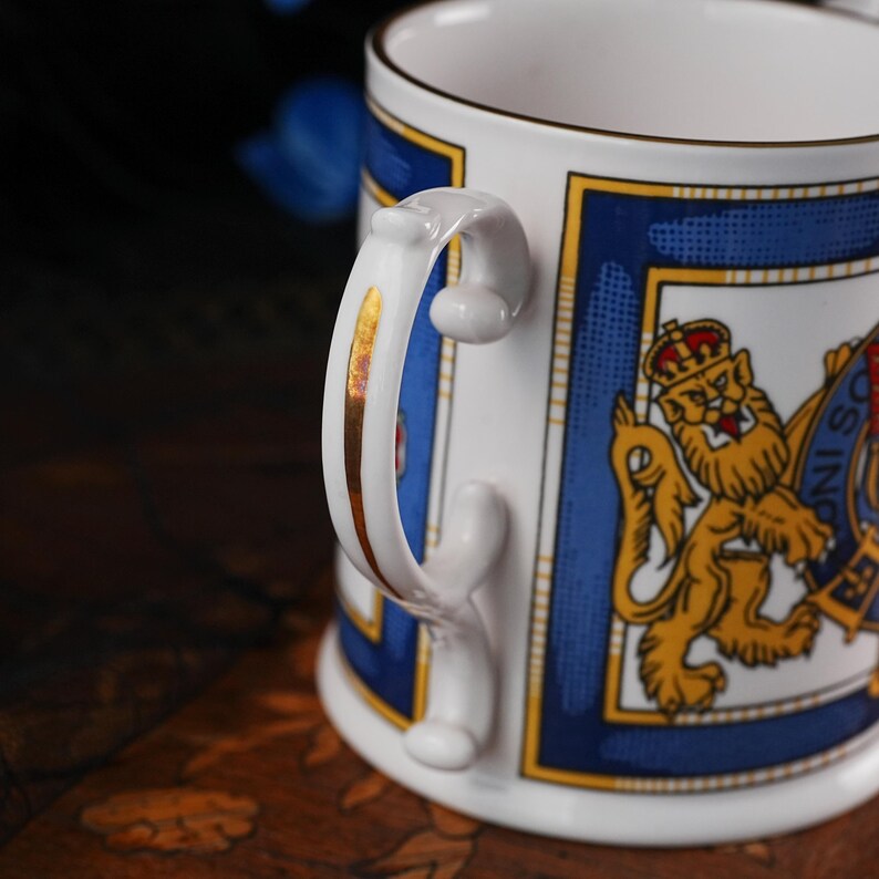 May include: A white ceramic mug featuring a gold-trimmed handle and a blue rectangular border. The design includes a detailed illustration of a golden lion wearing a crown, set against a white background with blue and yellow geometric accents.