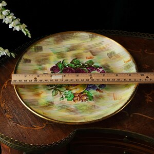 May include: A round decorative plate with a gold rim, featuring a hand-painted fruit design. The plate has a diameter of approximately 12 inches. The background has a yellow and green brushstroke pattern. The fruit includes grapes, berries, and a pear.
