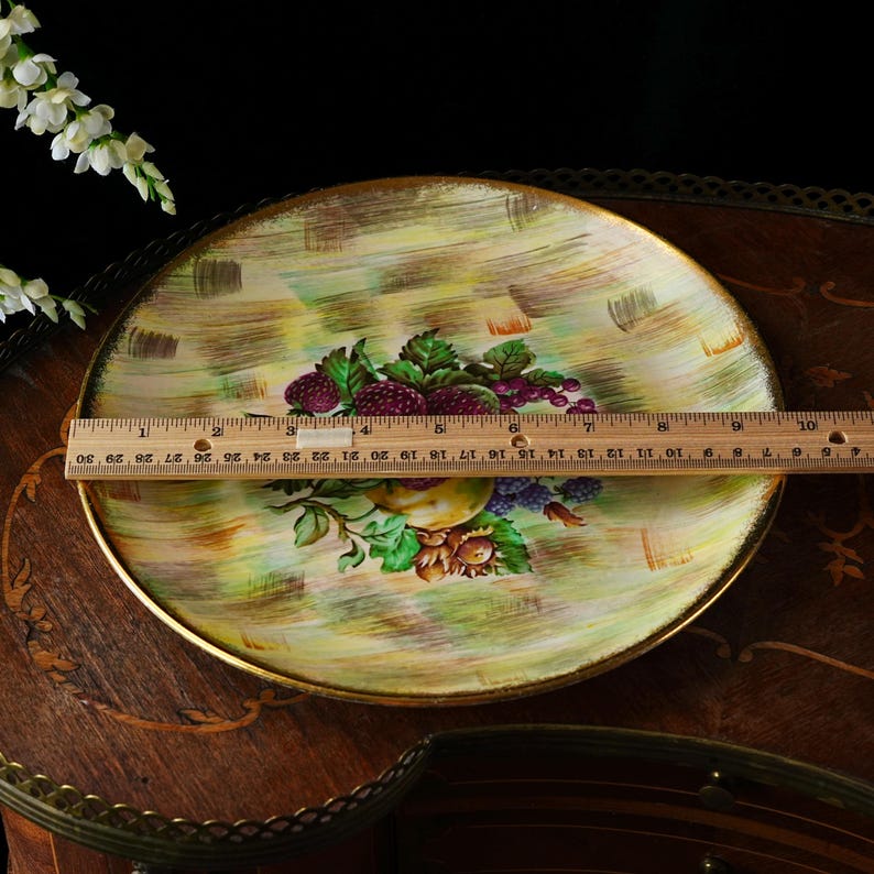 May include: A round decorative plate with a gold rim, featuring a hand-painted fruit design. The plate has a diameter of approximately 12 inches. The background has a yellow and green brushstroke pattern. The fruit includes grapes, berries, and a pear.