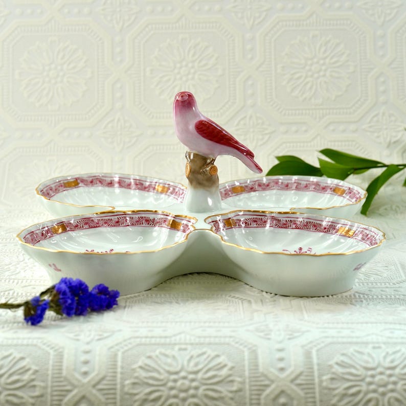 May include: A white porcelain serving dish with four compartments, each with a red and gold floral design. A pink and red bird figurine sits atop the dish. The dish is on a white surface.