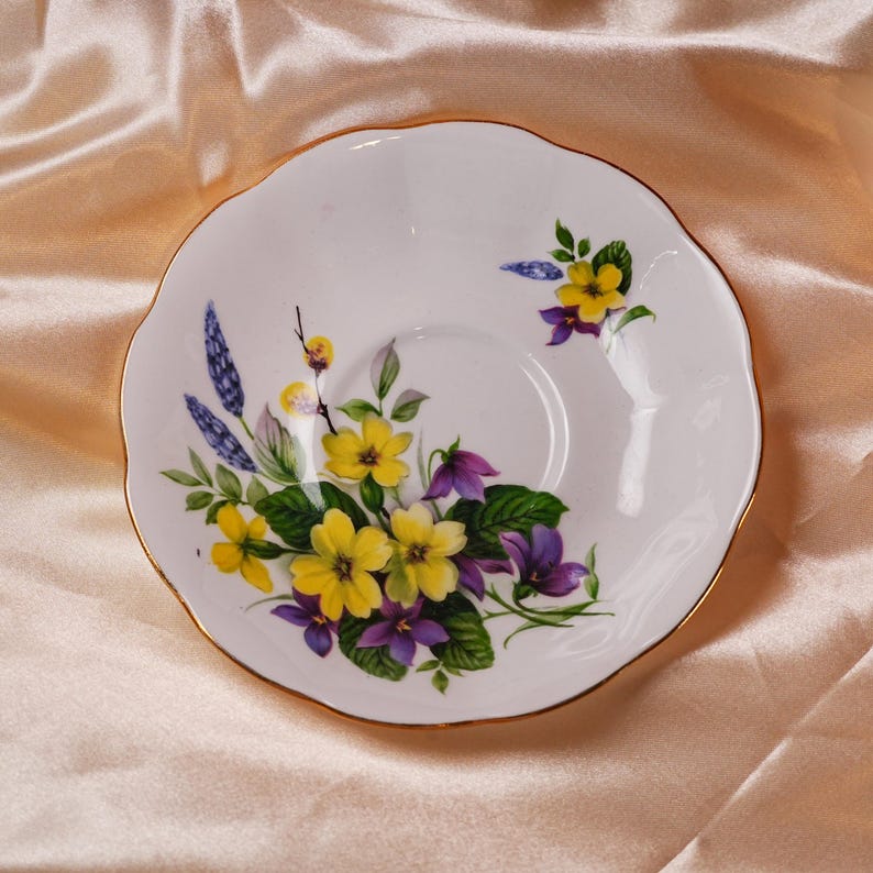 K&ouml;nnte beinhalten: Wei&szlig;e Porzellantasse mit gewelltem Rand und Goldrand. Die Untertasse ist mit einem Blumendesign mit gelben und lila Blumen, gr&uuml;nen Bl&auml;ttern und blauen Akzenten verziert.