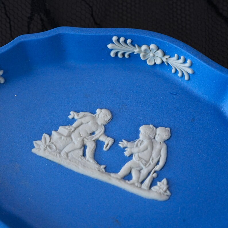 May include: A bright blue oval tray featuring white relief ornamentation. The centre displays a scene of three figures in white, while the scalloped rim is decorated with white floral and leaf motifs along the curved edge of the blue surface.