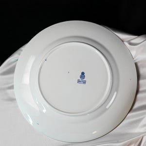May include: White ceramic plate with a blue emblem and text on the back. The plate has a wide rim and a slightly raised center. The emblem appears to be a crest or coat of arms. The plate is likely vintage or antique.