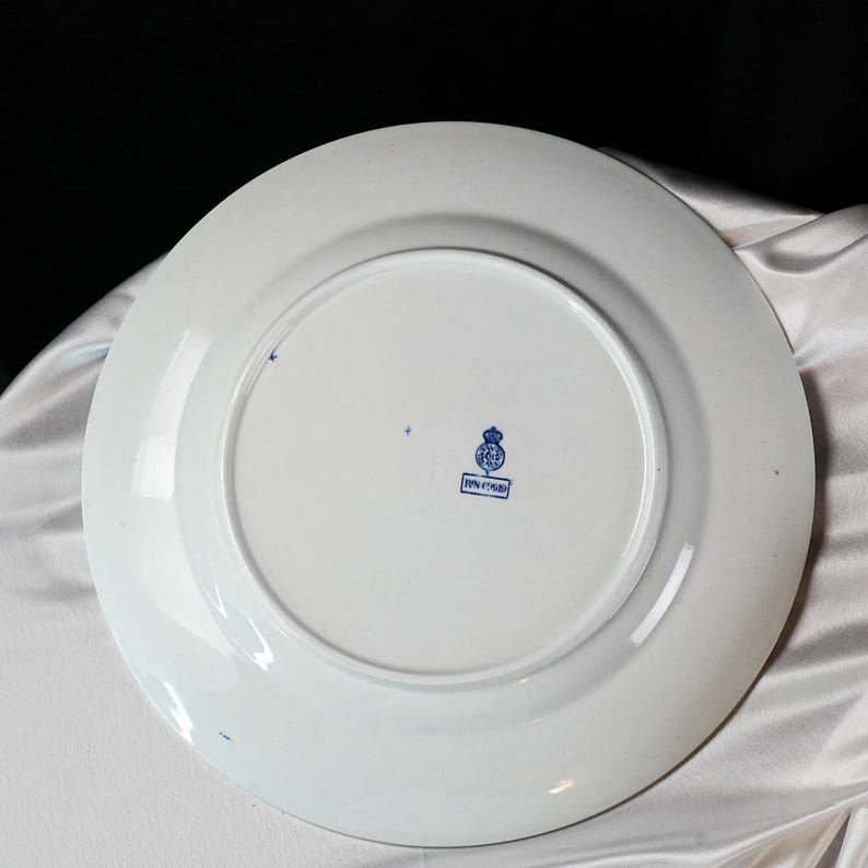 May include: White ceramic plate with a blue emblem and text on the back. The plate has a wide rim and a slightly raised center. The emblem appears to be a crest or coat of arms. The plate is likely vintage or antique.