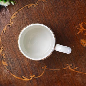 May include: A white ceramic mug with a handle, viewed from above, on a dark wooden surface. The mug has a gold-coloured rim and a white interior. The wooden surface features decorative inlaid patterns.
