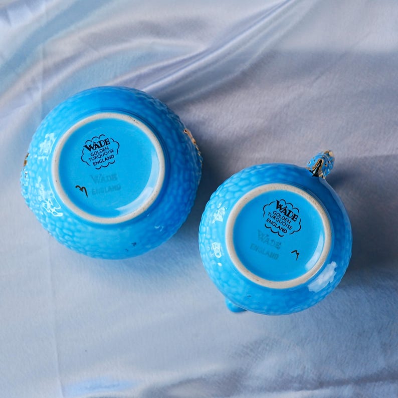 Op de afbeelding: Twee fel turquoise keramische vaten met een getextureerd, bobbelig oppervlak. De onderkant is voorzien van een zwarte stempel met de tekst Wade Golden Turquoise England, samen met een kleine handgeschreven markering en gouden accenten.