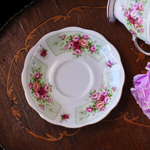 K&ouml;nnte beinhalten: Wei&szlig;e Porzellanuntertasse mit gewelltem Rand, verziert mit einem Blumenmuster aus rosa und roten Rosen auf hellgr&uuml;nem Hintergrund. Die Untertasse hat einen goldenen Rand. Eine passende Teetasse ist teilweise sichtbar.