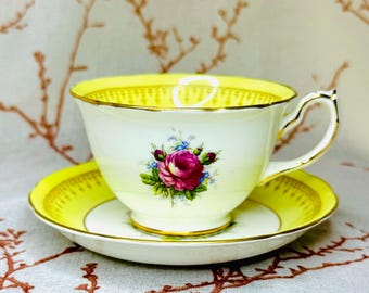 May include: A yellow and white teacup and saucer set with a floral design featuring a pink rose. The saucer has a gold trim.