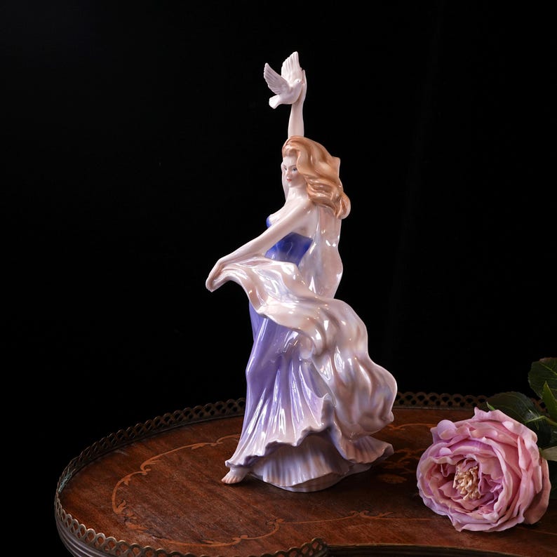 May include: A porcelain figurine of a woman in a flowing lavender gown, holding a white dove aloft. The figurine has blonde hair and is posed in a graceful dance. A pink rose rests on a wooden table.